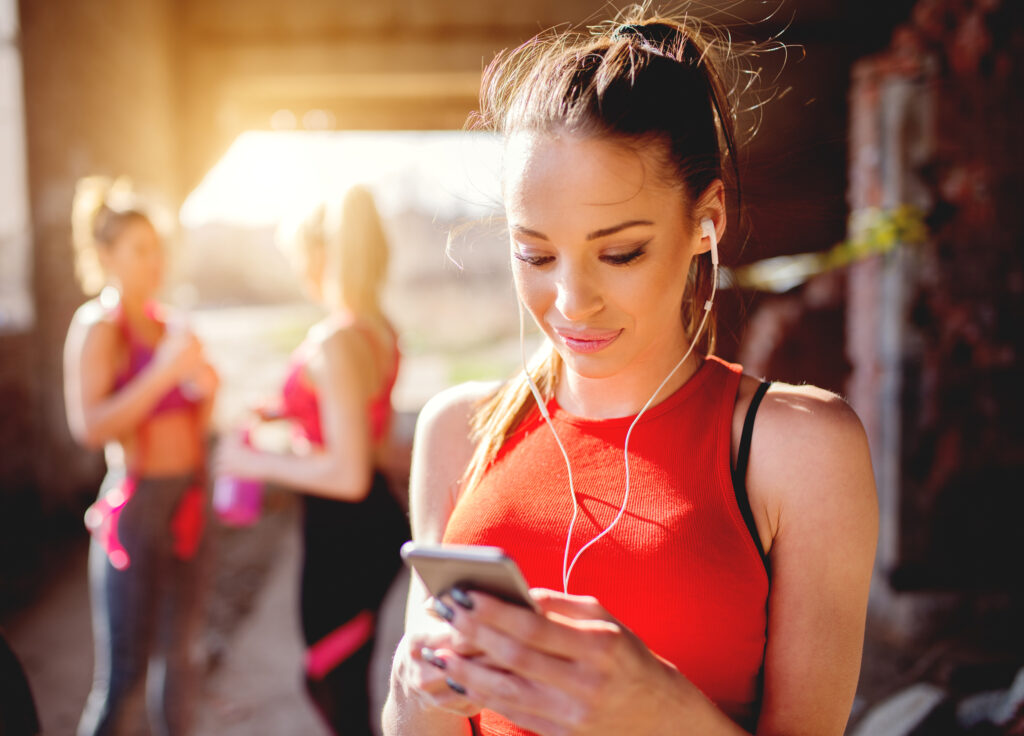 Young attractive fitness model listening music on smart phone charching positive energy before workout outside. Sun is shining and friends are behind preparing for training.